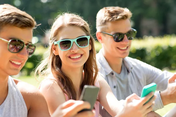 Amigos sonrientes con teléfonos inteligentes sentados en el parque —  Fotos de Stock