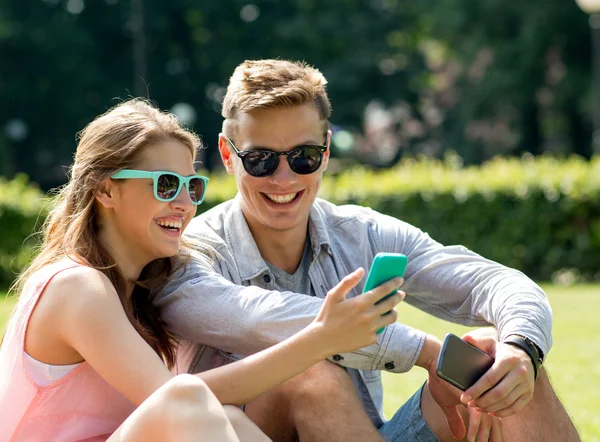 Amigos sonrientes con teléfonos inteligentes sentados en el parque — Foto de Stock