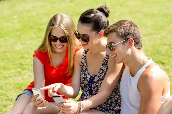 Amigos sonrientes con teléfonos inteligentes sentados en el parque — Foto de Stock