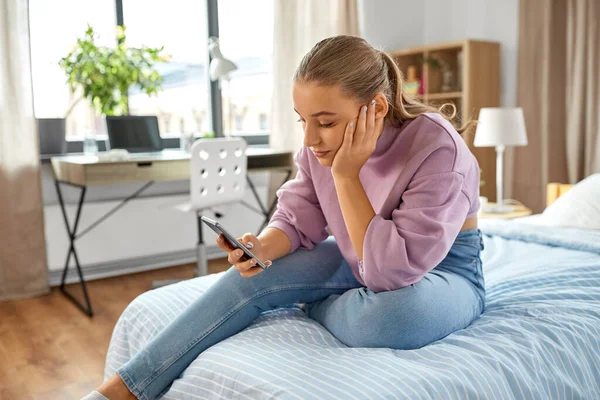 Λυπημένη κοπέλα με smartphone κάθεται στο κρεβάτι στο σπίτι — Φωτογραφία Αρχείου