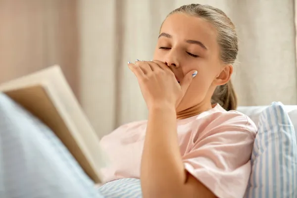 Yawning girl reading book in bed at home — Stock Photo, Image