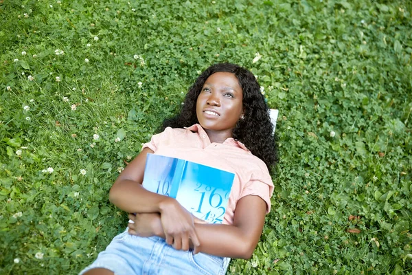 Afrikaans student meisje met leerboek liggend op gras — Stockfoto