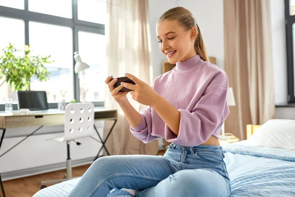 Happy girl with smartphone sitting on bed at home — Stock Photo, Image