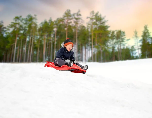 Menino feliz deslizando no trenó colina de neve no inverno — Fotografia de Stock