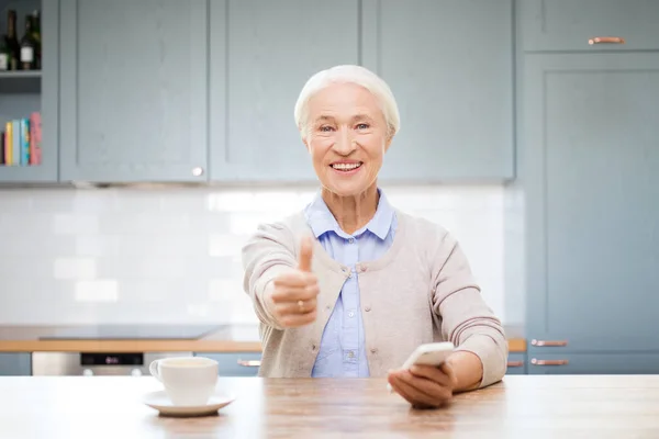 Женщина со смартфоном показывает большие пальцы на кухне — стоковое фото