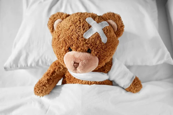Osito de peluche enfermo con pata vendada y cabeza en la cama —  Fotos de Stock