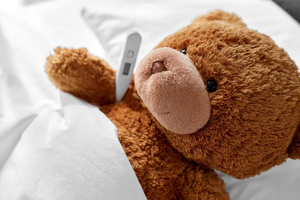 Больной плюшевый медвежонок с термометром в постели — стоковое фото