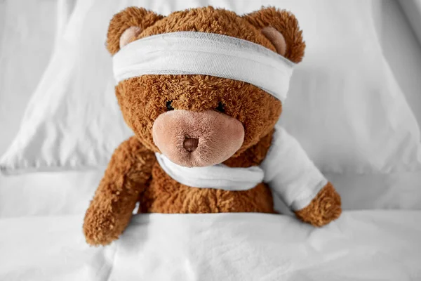 Ill teddy bear with bandaged paw and head in bed Stock Photo
