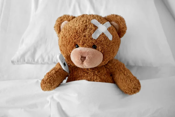 Ill teddy bear with patch and thermometer in bed — стоковое фото