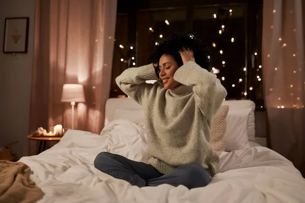 Mujer en suéter tocándose el pelo en la cama por la noche —  Fotos de Stock