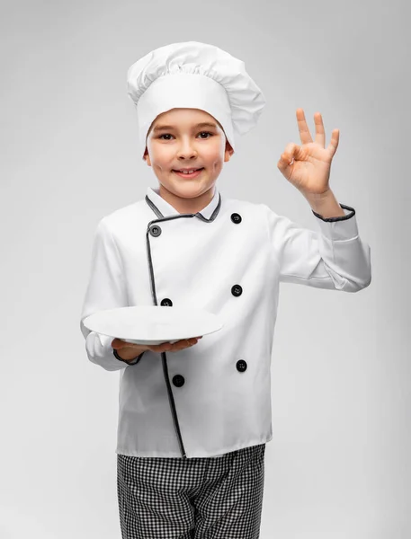 Smiling boy in chefs toque with plate showing ok — Stock Photo, Image