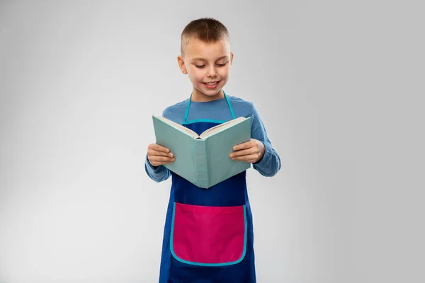 Niño sonriente en delantal leyendo libro de cocina — Foto de Stock