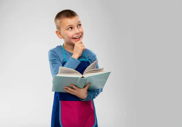 Niño en delantal leyendo libro de cocina y pensando —  Fotos de Stock