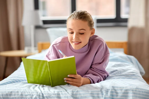 Uśmiechnięta dziewczyna pisząca do pamiętnika leżącego na łóżku w domu — Zdjęcie stockowe