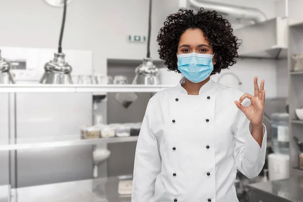 Vrouwelijke chef-kok in medisch masker tonen ok op keuken — Stockfoto