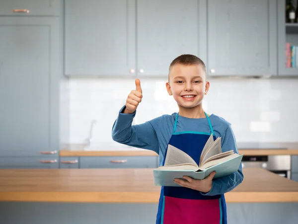 Menino em avental com livro de cozinha mostrando polegares para cima — Fotografia de Stock