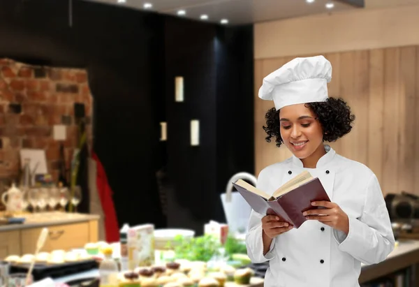 Szczęśliwy kobieta szef kuchni w białej toque czytania książki kucharskiej — Zdjęcie stockowe