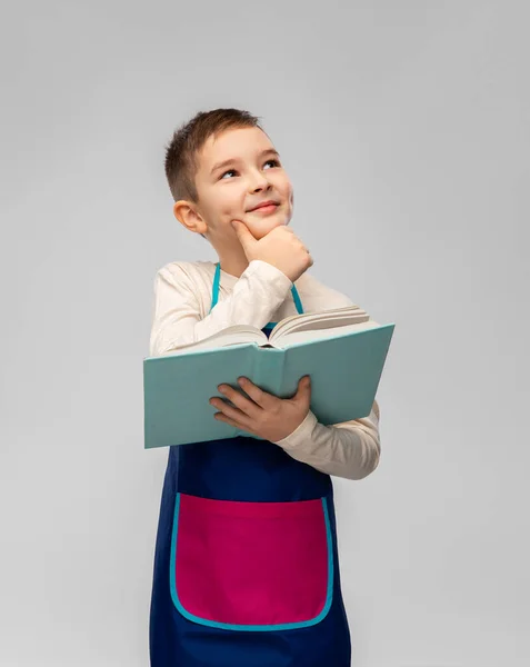 Malý chlapec v zástěře čtení kuchařky a přemýšlení — Stock fotografie