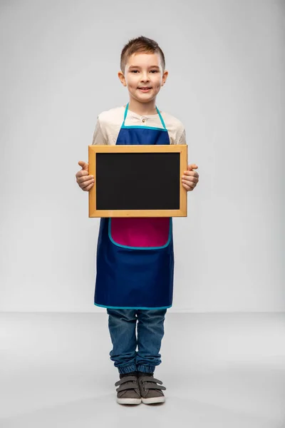 Smiling little boy in apron holding chalkboard — Stock Photo, Image