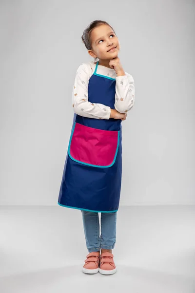 Thinking little girl in kitchen apron — стоковое фото