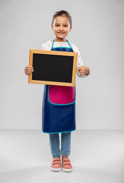 Niña sonriente en delantal sosteniendo pizarra — Foto de Stock