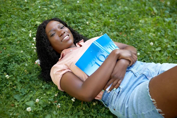 Студентка из Африки с учебником, лежащим на траве — стоковое фото
