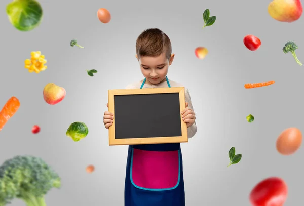 Little boy in apron holding chalkboard over food — Stock Photo, Image