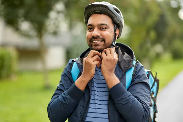 food shipping, safety and people concept - happy smiling delivery man with thermal insulated bag fastening his bicycle helmet on city street