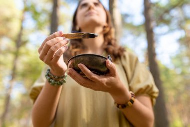 occult science and supernatural concept - close up of woman or witch with smoking palo santo stick and bowl performing magic ritual in forest clipart