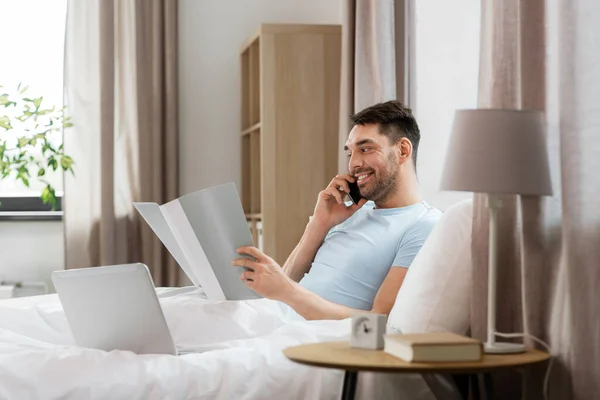 people, technology and remote job concept - man with folder and laptop computer calling on smartphone in bed at home bedroom