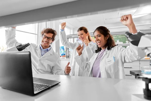 science, work and people concept - international group of happy smiling scientists with laptop celebrating success in laboratory