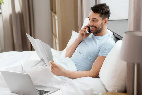 技術とリモートジョブの概念 家の寝室でベッドでスマートフォンを呼び出すフォルダとラップトップコンピュータを持つ男 — ストック写真