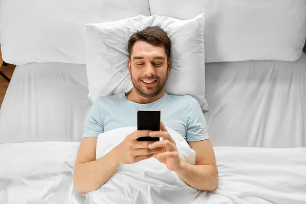 テクノロジーとインターネットと人々の概念 朝に自宅のベッドでスマートフォンにメッセージを送る笑顔の男 — ストック写真