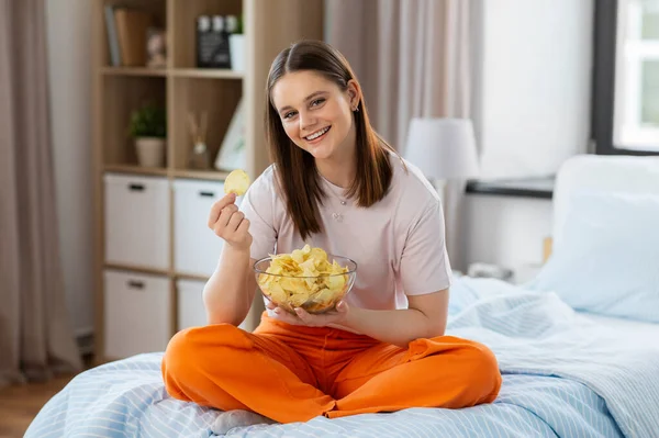 fast food and people concept - happy girl eating crisps sitting on bed at home