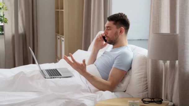 技術とリモートジョブの概念 家の寝室でベッドでスマートフォンを呼び出すフォルダとラップトップコンピュータを持つ男 — ストック動画