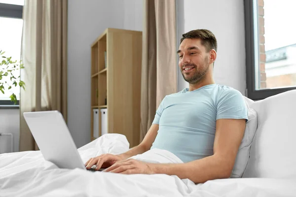 人と技術とリモートの仕事の概念 ラップトップコンピュータを持つ男が自宅のベッドルームに横たわっている — ストック写真