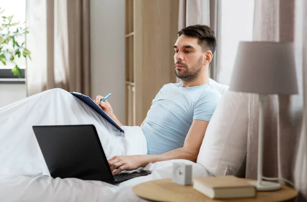 people, technology and remote job concept - man with laptop computer and clipboard working in bed at home bedroom