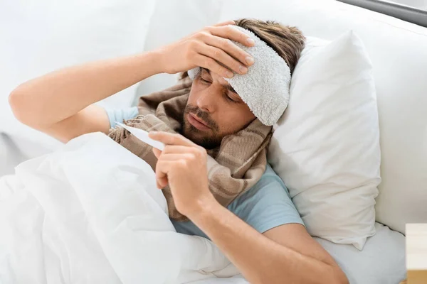 熱の概念 自宅でベッドに横たわって温度計によって彼の額の温度を測定する上で冷たい圧縮を持つ病人 — ストック写真
