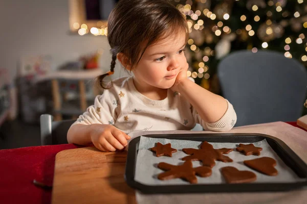 christmas, cooking and winter holidays concept - happy little baby girl with raw gingerbread cookies on baking tray at home
