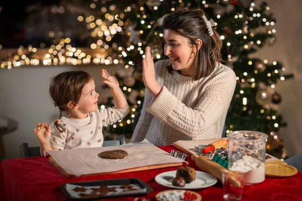 family, cooking and winter holidays concept - happy mother and baby daughter with rolling pin and dough for gingerbread cookies making high five gesture at home on christmas