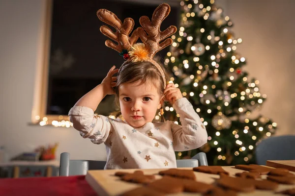 christmas, cooking and winter holidays concept - happy little baby girl with reindeer horns tiara and gingerbread cookies on table at home