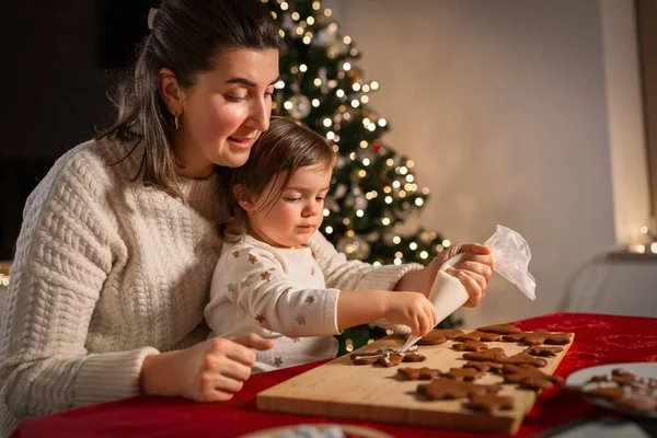 family, cooking and winter holidays concept - happy mother and baby daughter with icing in baking bag decorating gingerbread cookies at home on christmas