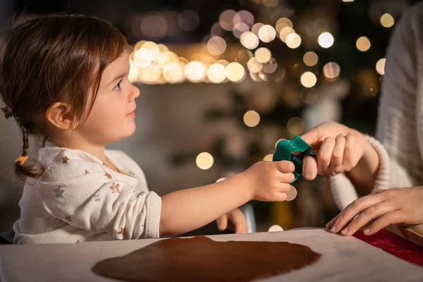 family, cooking and winter holidays concept - happy mother and baby daughter with mold making gingerbread cookies from dough at home on christmas