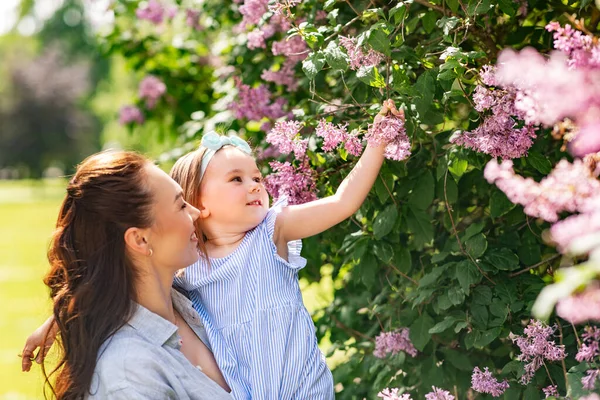 family, motherhood and people concept - happy mother with little daughter touching lilac flowers at summer park or garden