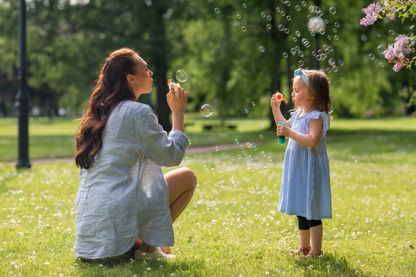 family, motherhood and people concept - happy mother with little daughter blowing soap bubbles at summer park or garden