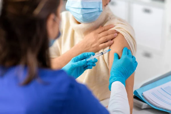 medicine, health and vaccination concept - close up of doctor or nurse with syringe making vaccine or drug injection to senior woman in mask at hospital