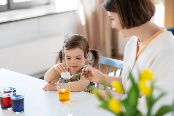 holidays, family and people concept - happy mother and little baby daughter dyeing easter eggs with colors at home