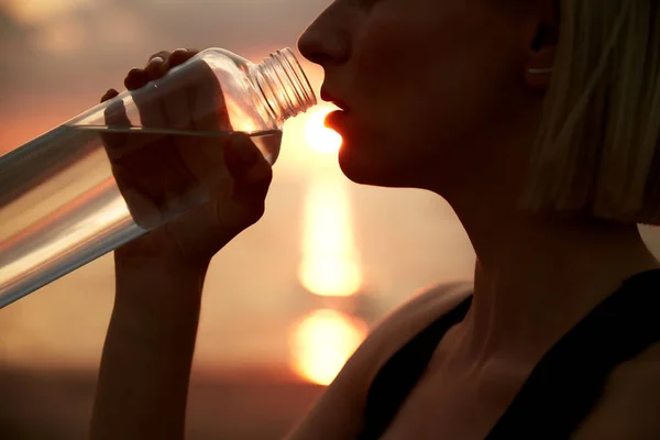 fitness, sport, and healthy lifestyle concept - close up of woman drinking water from bottle on beach over sunset