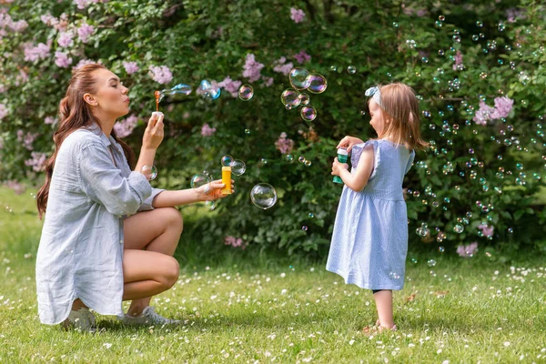 family, motherhood and people concept - happy mother with little daughter blowing soap bubbles at summer park or garden
