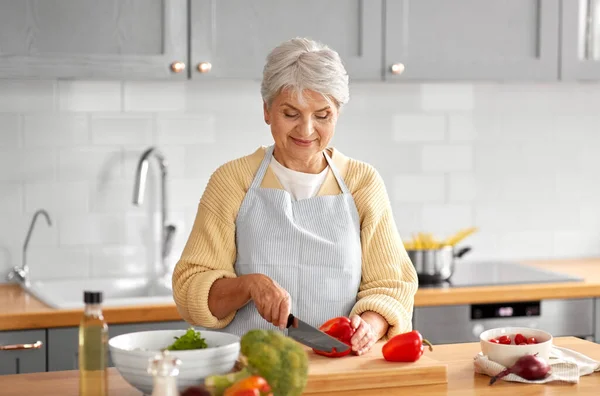 健康的な食事 料理のコンセプト 家庭でキッチンで赤唐辛子を切りナイフで幸せな笑顔のシニア女性 — ストック写真